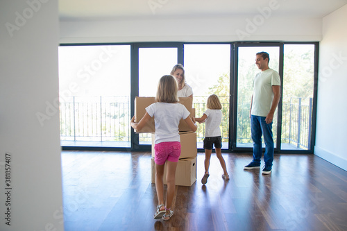 Caucasian man, woman and kids with stacked carton boxes standing in empty room of new apartment and smiling. Back view of girl carrying cardboard box. Mortgage, relocation and moving day concept