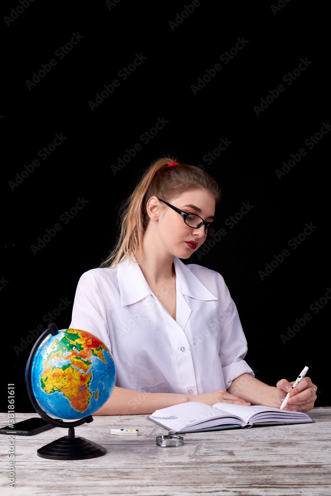 Yong female teacher is sitting at the desk. Beautiful teacher is writing on black background. Yong female teacher is sitting at the desk. Beautiful teacher is writing on black background.
