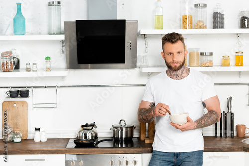 young bearded man in white t-shirt looking at camera while holding bowl with breakfast in kitchen