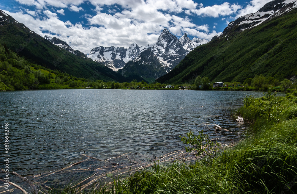 Mountain lake. Reed floats in the water, green and rocky snowy mountains surround the plain, blue sky. Caucasus., Russia