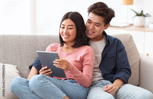 Cheerful Asian Couple Using Digital Tablet Sitting On Couch Indoors
