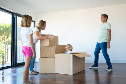 Happy parents and two daughters having fun while unpacking things in their new empty flat. Girl sticking out from carton box. Real estate purchase concept