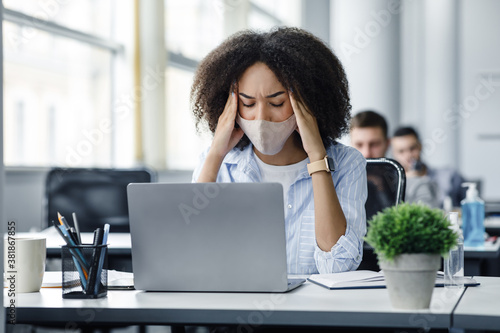 Overwork, illness and disease symptoms in workers. African american woman in protective mask suffers from headache, sitting at table with antiseptic, laptop in office interior