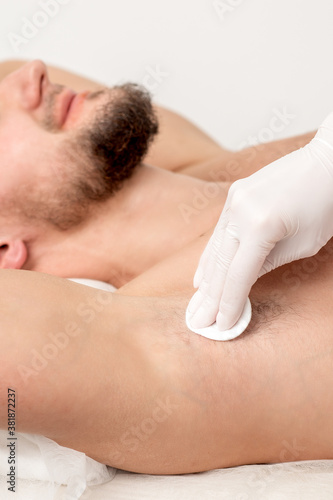 Hands of depilation master in white rubber gloves wiping male armpit with a cotton pad
