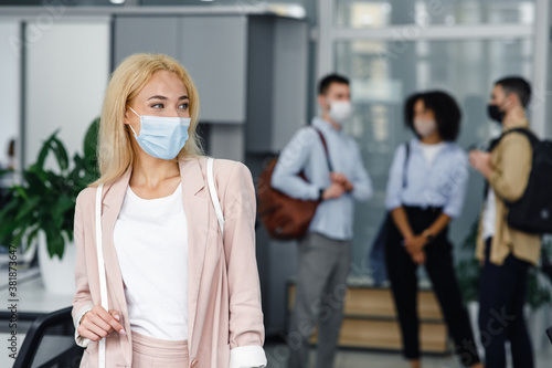 Return to work after lockdown and new normal. Woman in protective mask looks to the side in corridor of modern office with blurred colleagues