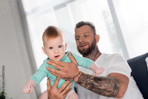 selective focus of baby boy looking at camera in hands of young father