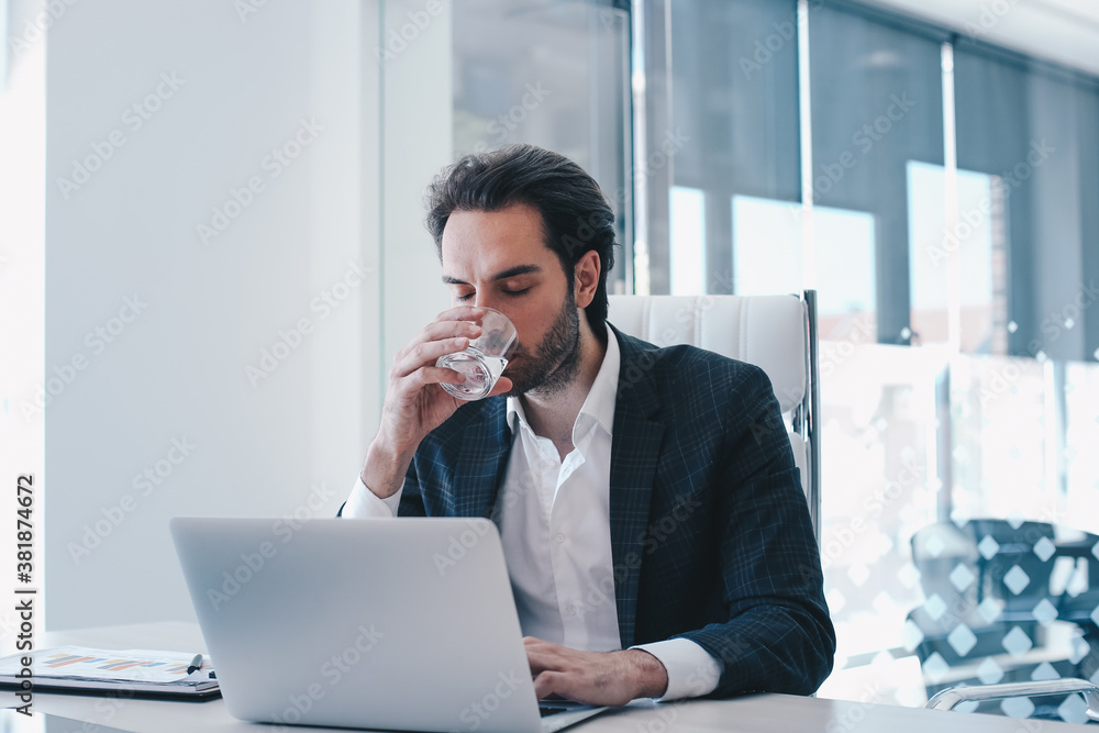 Young handsome unshaven businessman in suit sitting in his office, drinking water and finishing his report. Young handsome unshaven businessman in suit sitting in his office, drinking water and finishing his report.