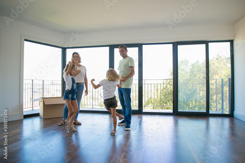 Joyful kids dancing and jumping in empty room. Happy parents looking at daughters and smiling. Cheerful family moving in big new apartment or house. Mortgage, relocation and moving day concept © Mangostar