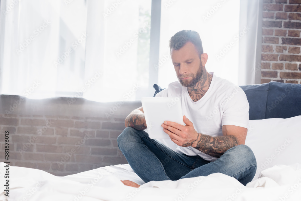 tattooed man in white t-shirt and jeans using digital tablet while sitting in bed with crossed legs tattooed man in white t-shirt and jeans using digital tablet while sitting in bed with crossed legs