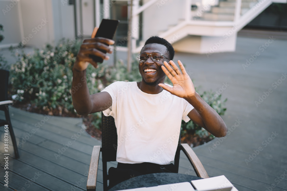 Cheerful dark skinned african american male blogger using smartphone camera for making vlog video sitting on cafe terrace, cheerful african american man waving during mobile phone online call Cheerful dark skinned african american male blogger using smartphone camera for making vlog video sitting on cafe terrace, cheerful african american man waving during mobile phone online call