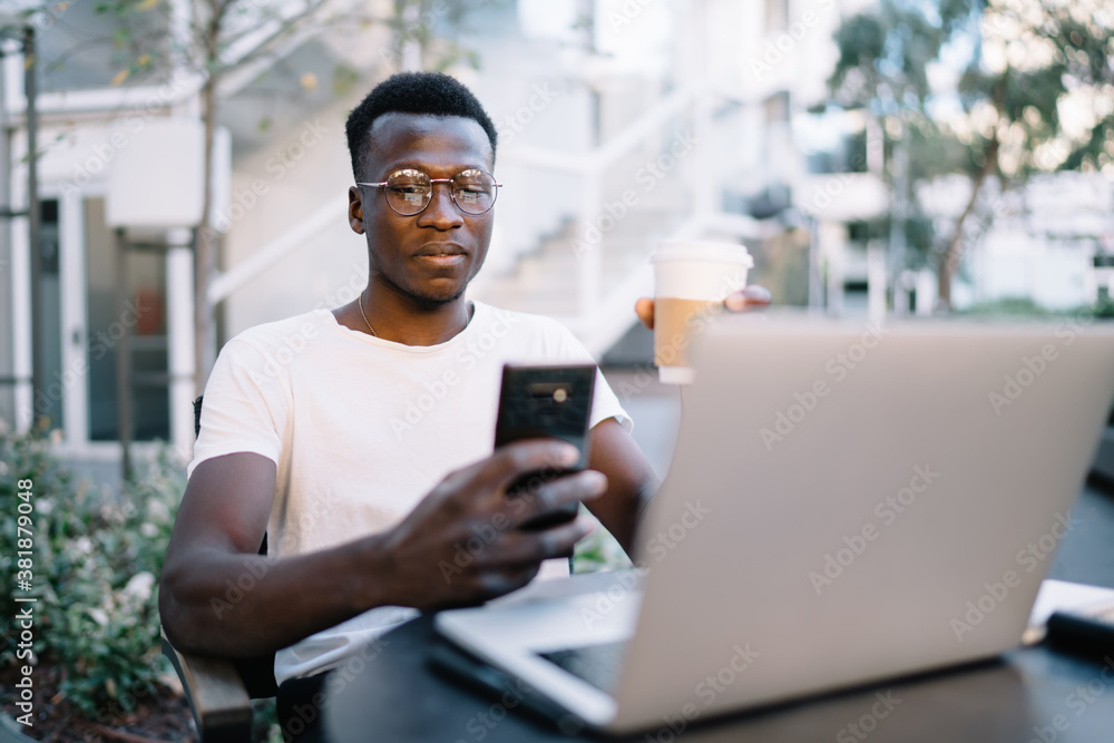Pensive prosperous male freelancer sitting near laptop computer for remote job using smartphone for checking notification, smiling dark skinned man blogger using mobile phone share media content Pensive prosperous male freelancer sitting near laptop computer for remote job using smartphone for checking notification, smiling dark skinned man blogger using mobile phone share media content