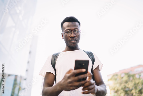 Pensive african american male blogger share multimedia content online standing with mobile phone in city, pensive dark skinned hipster guy in casual wear and eyeglasses dialing number on smartphone © BullRun