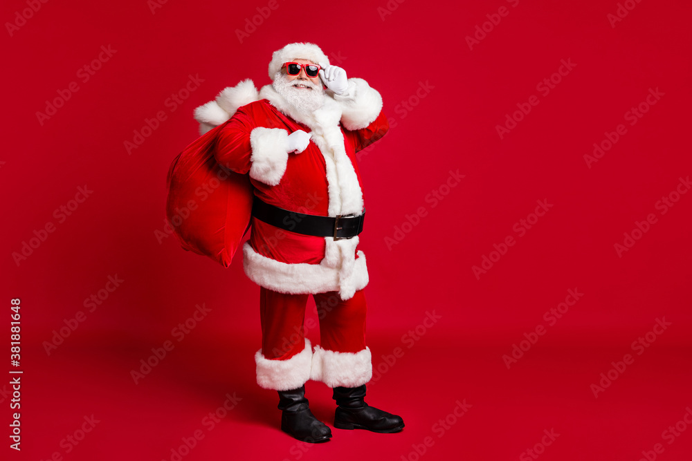 Full length photo of retired old man grey beard hold glasses bear sack presents good children wear santa costume gloves coat belt sunglass headwear black boots isolated red color background Full length photo of retired old man grey beard hold glasses bear sack presents good children wear santa costume gloves coat belt sunglass headwear black boots isolated red color background