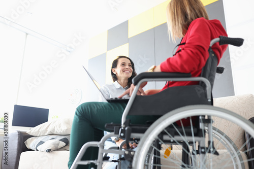 Disabled woman in wheelchair communicates with smiling friend with laptop on her lap. Psychological assistance to people with disabilities concept.