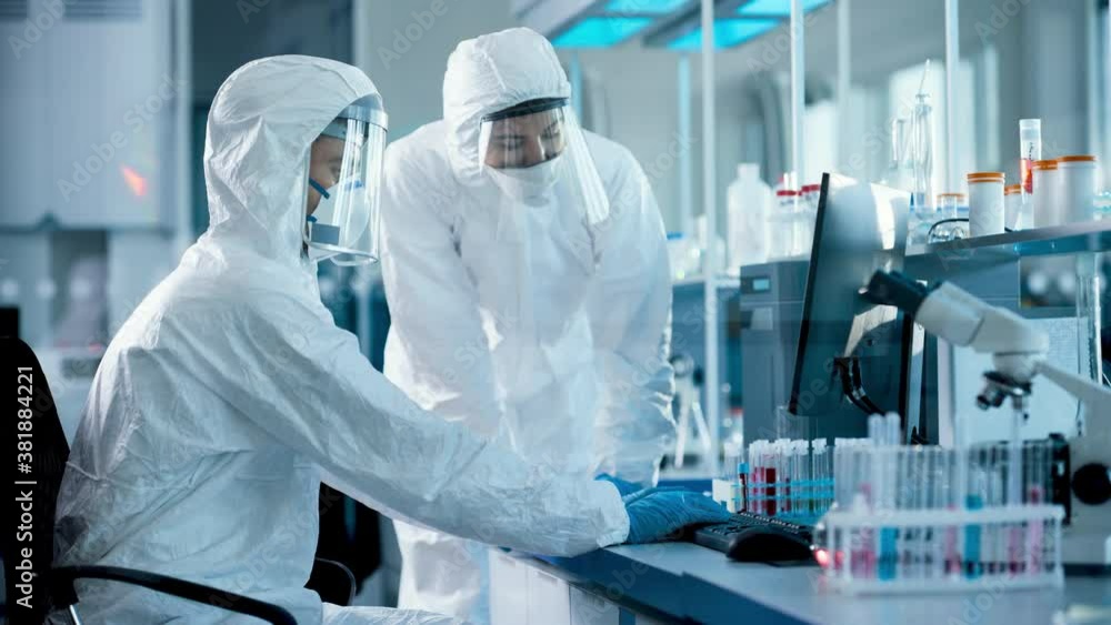 Medical Laboratory: Team of Microbiology Scientists Wearing Sterile Coveralls, Face Shields and Masks Talk, Use Computer to Analyse Test Tube Blood Samples and Develop Vaccine, Drugs and Antibiotics Medical Laboratory: Team of Microbiology Scientists Wearing Sterile Coveralls, Face Shields and Masks Talk, Use Computer to Analyse Test Tube Blood Samples and Develop Vaccine, Drugs and Antibiotics
