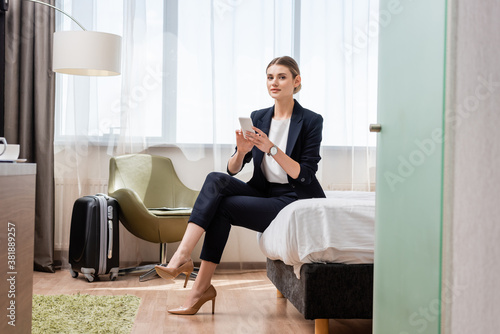 young businesswoman in suit holding smartphone while sitting on bed neat travel bag in hotel room