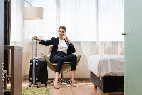selective focus of pleased businesswoman in suit talking on smartphone and sitting in armchair near baggage in hotel