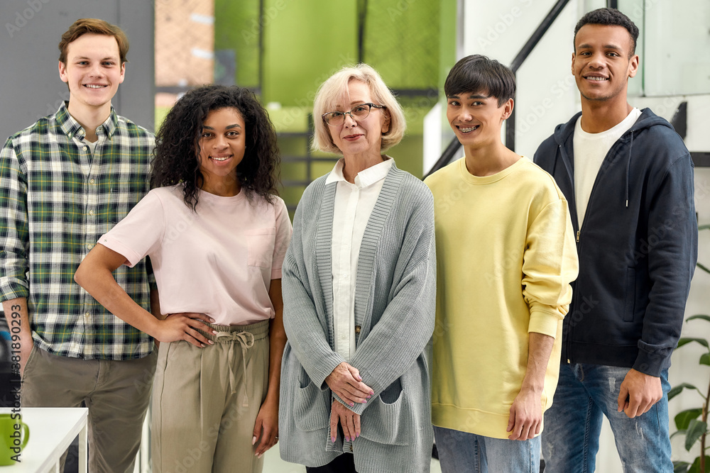 Happy diverse team of employees looking at camera while posing with senior female intern, standing together in the office, Age diversity in the workplace Happy diverse team of employees looking at camera while posing with senior female intern, standing together in the office, Age diversity in the workplace