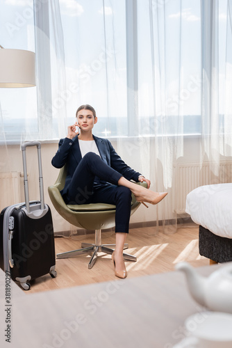 young businesswoman in suit talking on smartphone while sitting in armchair near luggage in hotel