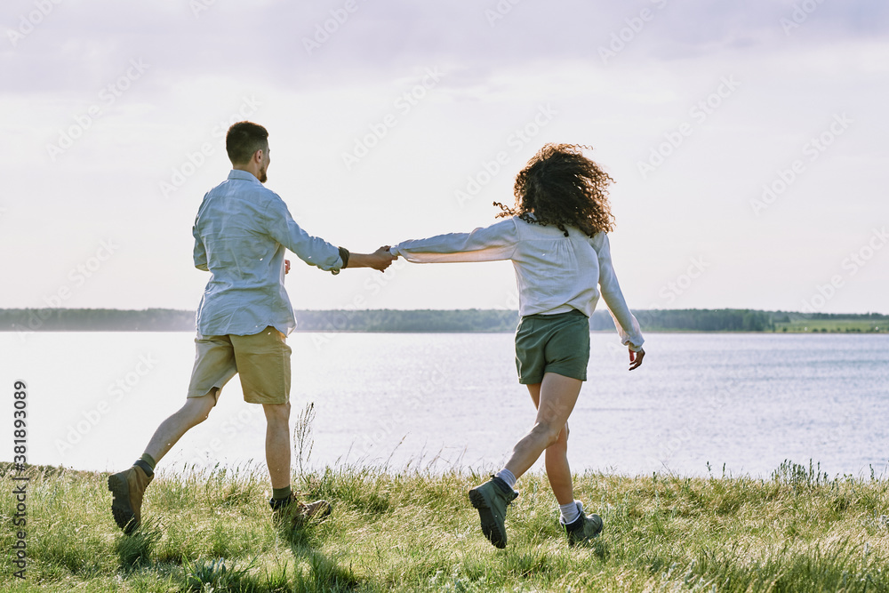 Rear view of affectionate young couple of hikers holding hands and running to lake while enjoying vacation Rear view of affectionate young couple of hikers holding hands and running to lake while enjoying vacation