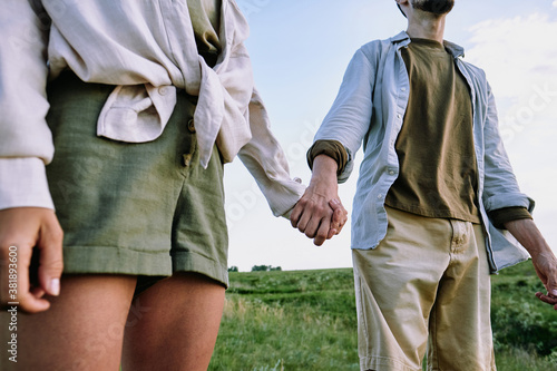 Close-up of unrecognizable travelling couple in casual shirts holding hands in hills while standing in field