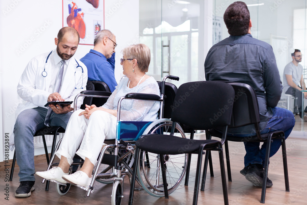 Doctor holding tablet pc with x-ray while explaining diagnosis to disabled elderly woman in wheelchair. Handicapped patient in hospital waiting area. Man in examination room. Doctor holding tablet pc with x-ray while explaining diagnosis to disabled elderly woman in wheelchair. Handicapped patient in hospital waiting area. Man in examination room.