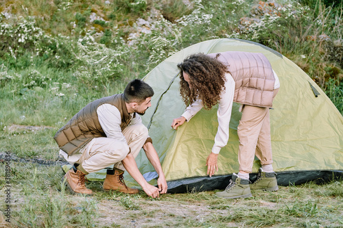 Beautiful young couple in vests setting up tent while preparing for camping outside