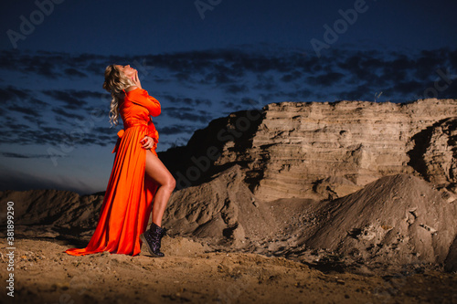 Woman in orange dress posing near the sand hill at the background of sunset