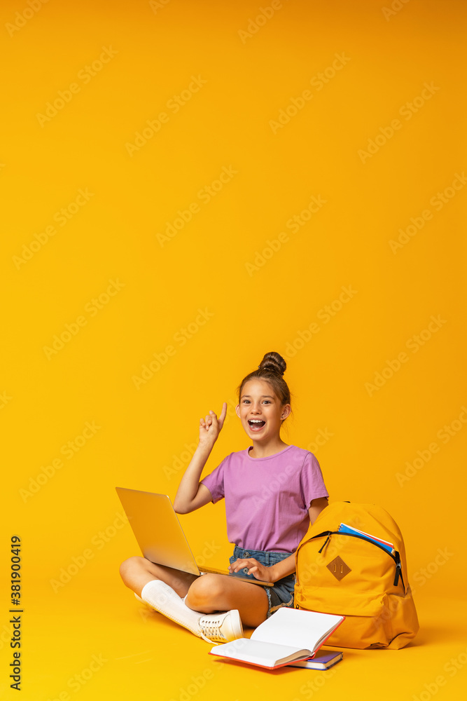 happy schoolgirl with backpack, laptop and books is sitting on yellow background