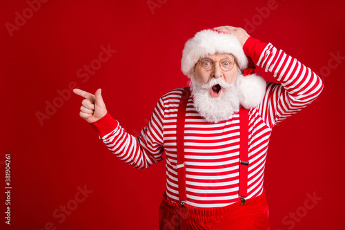 Photo of retired old man grey beard direct finger empty space open mouth hand head amazing decor wear santa costume suspenders spectacles striped shirt headwear isolated red color background © deagreez
