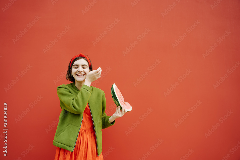 happy brunette girl Bob cut hair style red dress and green jacket holding a piece of watermelon and laughing. Red wall background. Having fun youth concept. hair accessories, mole on the face Teenager