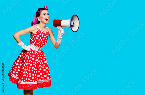 Violet red haired woman using mega phone, shouting something. Girl in pinup style dress in polka dot isolated over aqua blue color background. Retro fashion vintage studio concept. Copy space for text