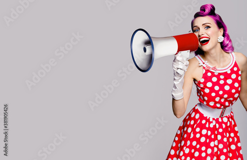 Purple head woman using mega phone, shouting something. Pinup girl in red polka dot dress. Retro vintage studio concept. Grey color background. Copy space place for some text.