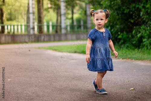 Little smiling cute dark-haired girl 1-3 in a denim dress walks along the road in the park on the backdrop of greenery.