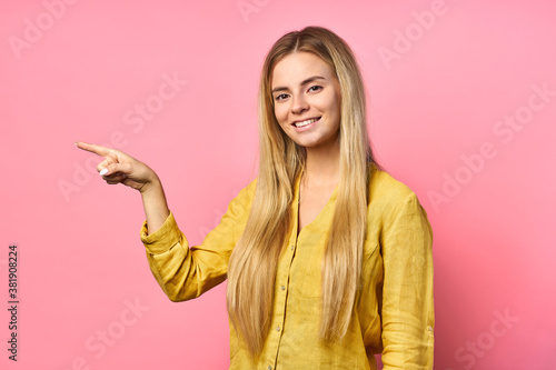 Portrait of a young blonde positive woman with a cheerful facial expression rejoices. shows a finger to the side, isolated on a pink background. Beautiful woman indoors.