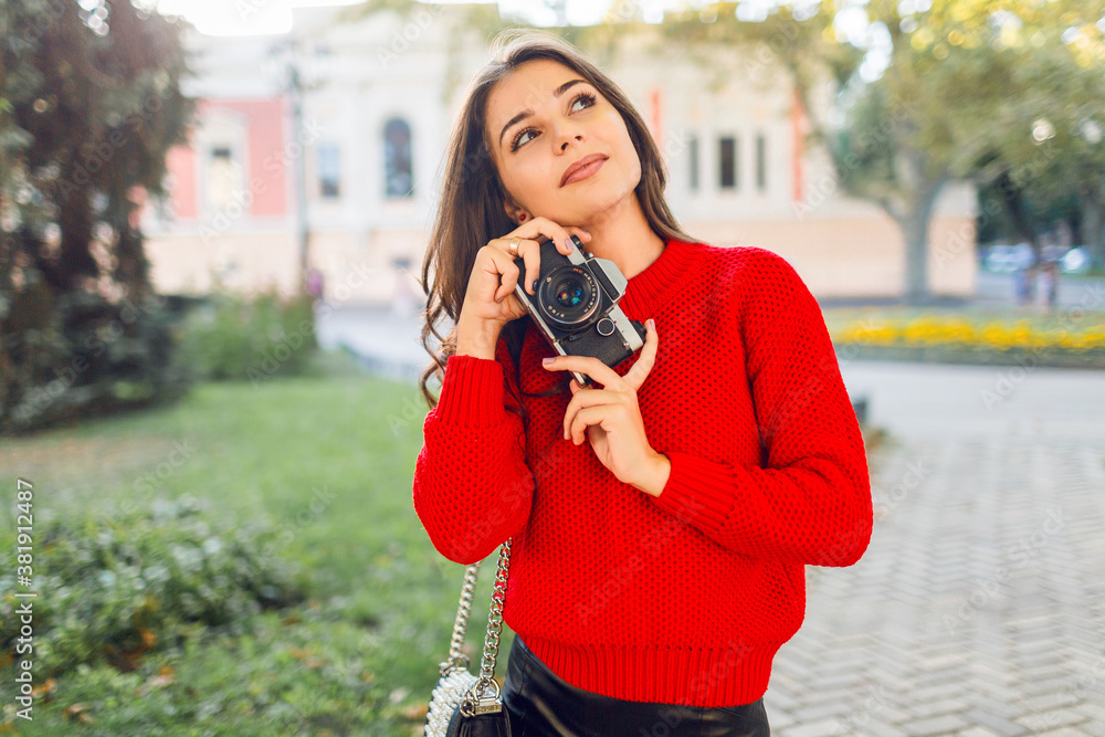 Sunny lifestyle image of pretty  brunette girl in red casual pullover and skirt making pictures by photo camera in sunny park. Walking in city garden and looking sightseens .