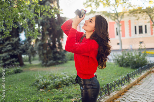 Sunny lifestyle image of pretty  brunette girl in red casual pullover and skirt making pictures by photo camera in sunny park. Walking in city garden and looking sightseens . photo