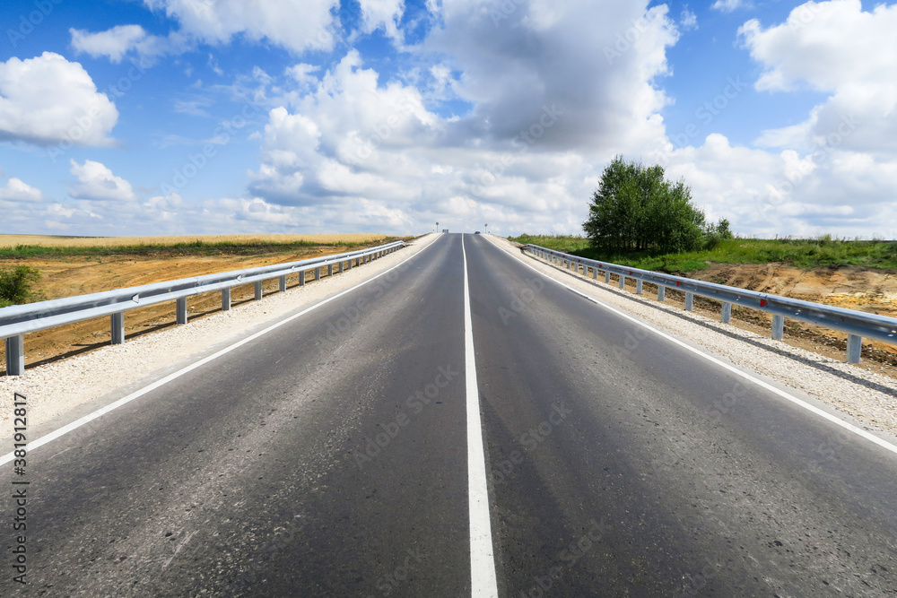 New asphalt road through the field. Close-up of an empty asphalt road. Clear horizon, blue sky and low-slung road. Green grass and yellow sunflowers on the roadside.