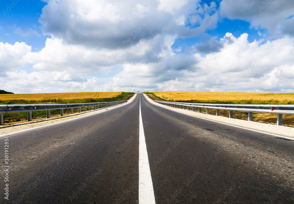 New asphalt road through the field. Close-up of an empty asphalt road. Clear horizon, blue sky and low-slung road. Green grass and yellow sunflowers on the roadside. New asphalt road through the field. Close-up of an empty asphalt road. Clear horizon, blue sky and low-slung road. Green grass and yellow sunflowers on the roadside.