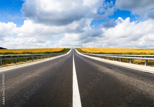 New asphalt road through the field. Close-up of an empty asphalt road. Clear horizon, blue sky and low-slung road. Green grass and yellow sunflowers on the roadside.