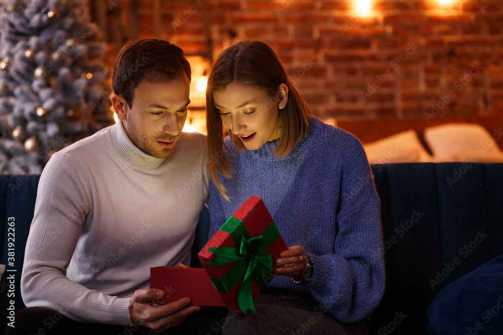 Handsome man presenting a gift to his beautiful wife and smiling. Beautiful young couple at home enjoying spending time together. Winter holidays, Christmas celebrations, New Year concept. Handsome man presenting a gift to his beautiful wife and smiling. Beautiful young couple at home enjoying spending time together. Winter holidays, Christmas celebrations, New Year concept.