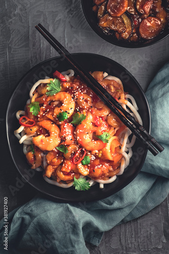 Flat lay of Japanese udon noodles fried with sweet and sour shrimps sauce with hot pepper in Asian style, black dishware, concrete background, dark mood