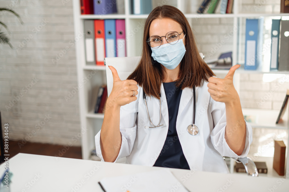 Female doctor wearing mask on her face sitting at the desk, looks at camera and shows thumbs up. Preventive measures during pandemic