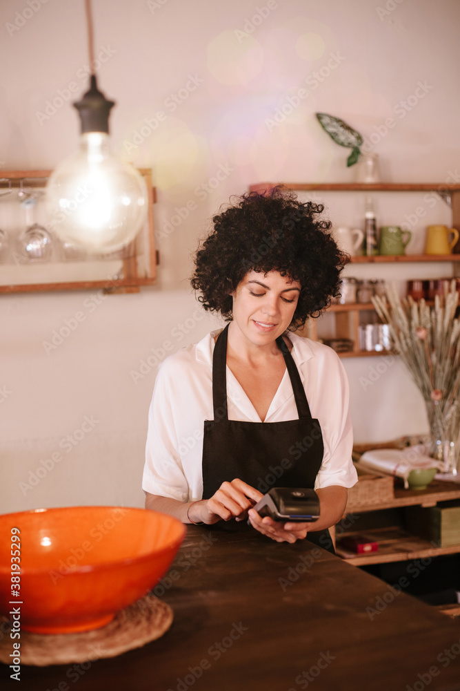 Beautiful female cafe owner standing with a card terminal. Woman with apron standing behind the bar counter of a modern restaurant. Concept small business owner, young entrepreneur. Beautiful female cafe owner standing with a card terminal. Woman with apron standing behind the bar counter of a modern restaurant. Concept small business owner, young entrepreneur.