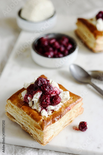 Puff pastry squares with ricotta and cherry sauce on white marble tray. Baked small square cakes topped with cheese and berry sauce
