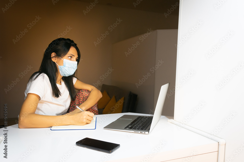 Young attractive caucasian woman  is holding pen and writing notes in front of laptop in cosy room in the background.