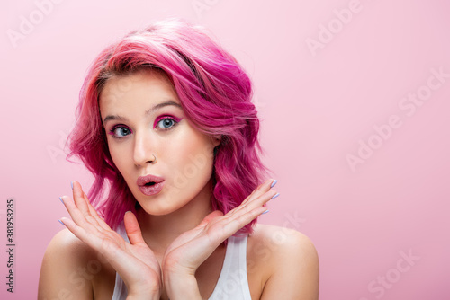 surprised young woman with colorful hair and makeup posing with hands near face isolated on pink