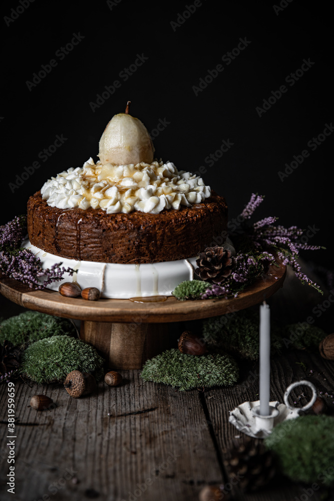 Homemade Carrot cake with ricotta-honey  frosting and whole pear on wooden cake stand surrounded with moss, acorns, pines and with little white candle on foreground on black background.