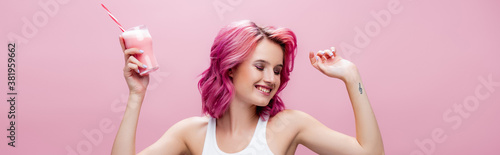 young woman with colorful hair holding strawberry milkshake in glass with drinking straw and dancing isolated on pink, panoramic shot