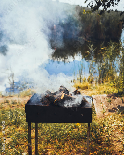 Metal brazier with fire wood and big smoke close up at nature background  near lake with trees and green grass  grill barbeque  picnic  fishing summer weekend concept outdoors 
